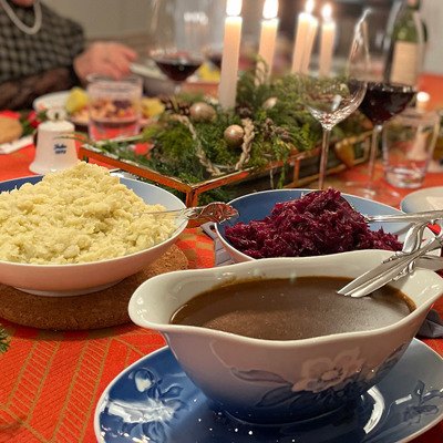 Christmas Eve with traditional holiday dishes and a table set with Bing & Groendahl Christmas Rose service. Serving dishes with red cabbage, potatoes, and gravy are placed on a red tablecloth surrounded by Christmas decorations and candlelight.