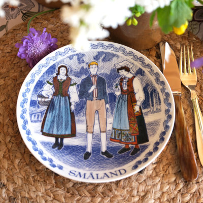 Ravn plate with a motif of folk costumes from Småland in Sweden, used as a dinner plate in a table setting with a woven placemat, flowers, and golden cutlery.