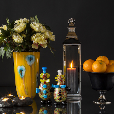 Two glass clown figures in front of yellow glass vase, lantern, and fruit bowl