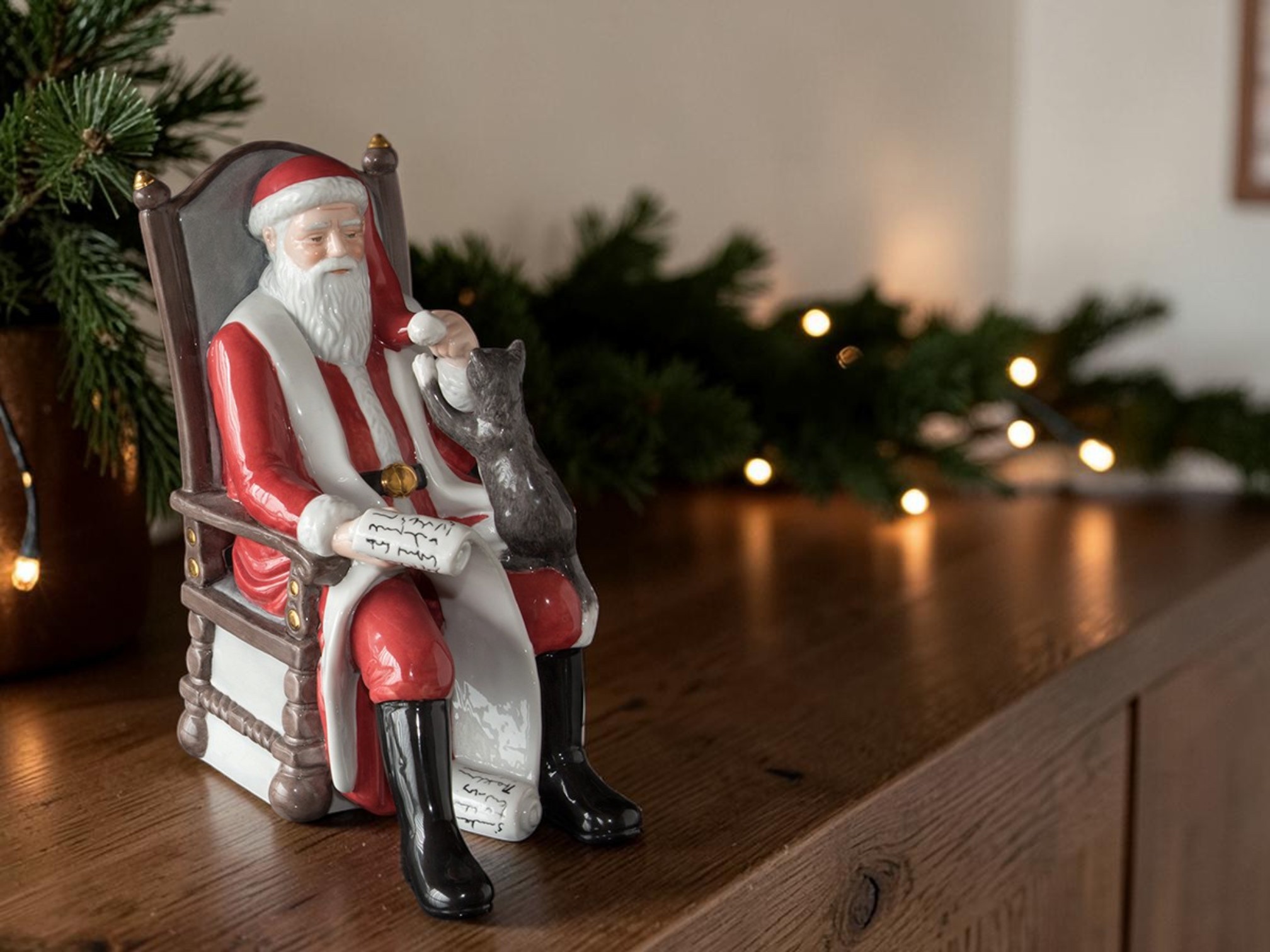 Royal Copenhagen Santa Claus figure in a rocking chair with a small cat on the lap, placed on a wooden dresser with fir branches and Christmas lights in the background.