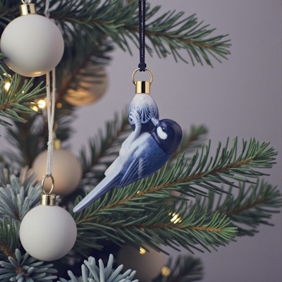 Royal Copenhagen ornament 2021 with blue tit hangs on the Christmas tree among green branches, light Christmas balls, and warm Christmas lights.