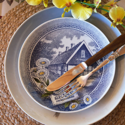 Table setting with a Ravn landscape plate used as a starter plate on top of a blue service. A woven placemat and flowers in yellow tones complete the personal look.