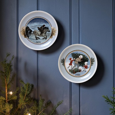 Two Tettau Christmas plates with winter motifs hanging on a dark blue wall above fir and warm Christmas lights.