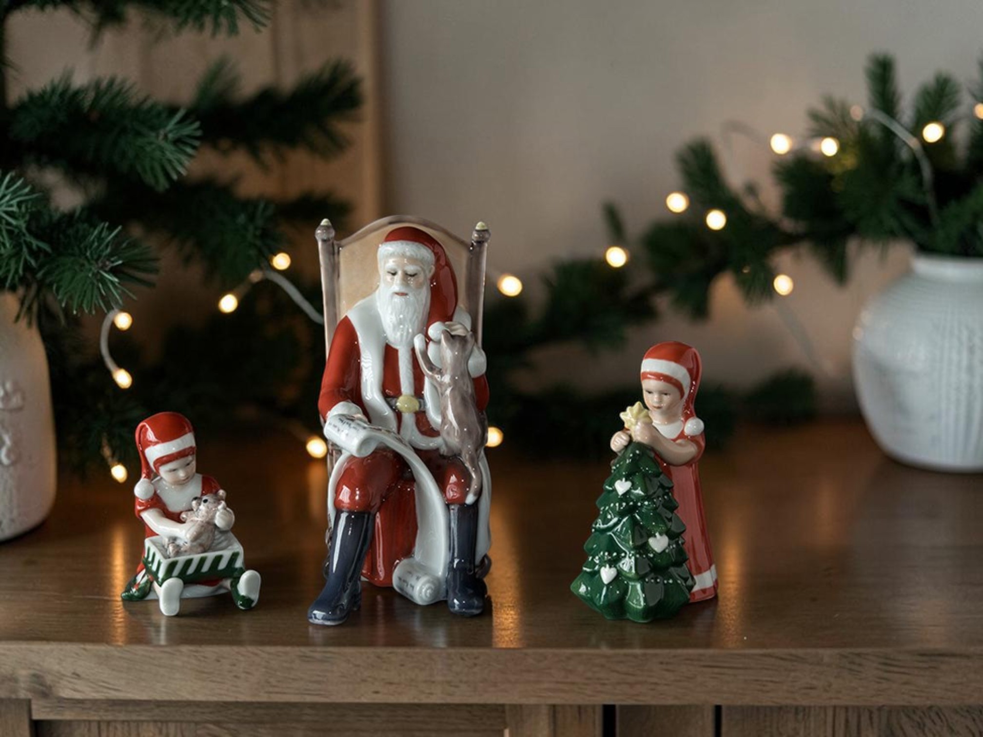 Royal Copenhagen Santa Claus in a rocking chair with two small figures from the Christmas with Else series, surrounded by fir branches and Christmas lights.