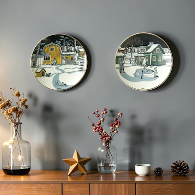 Two Arabia Christmas plates with colorful winter motifs hang on a gray wall above a sideboard with branches, cones, and Christmas decorations.
